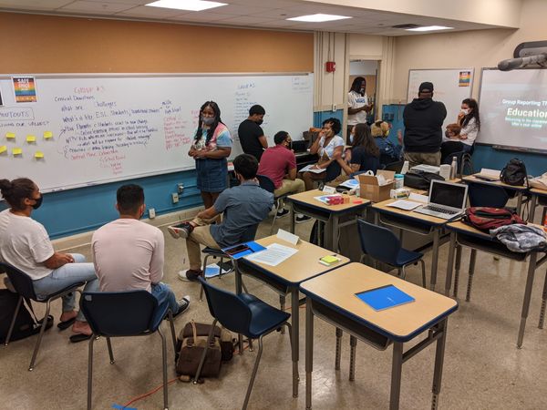 Students sit in a classroom with desks and computers in front of a white board filled with notes.